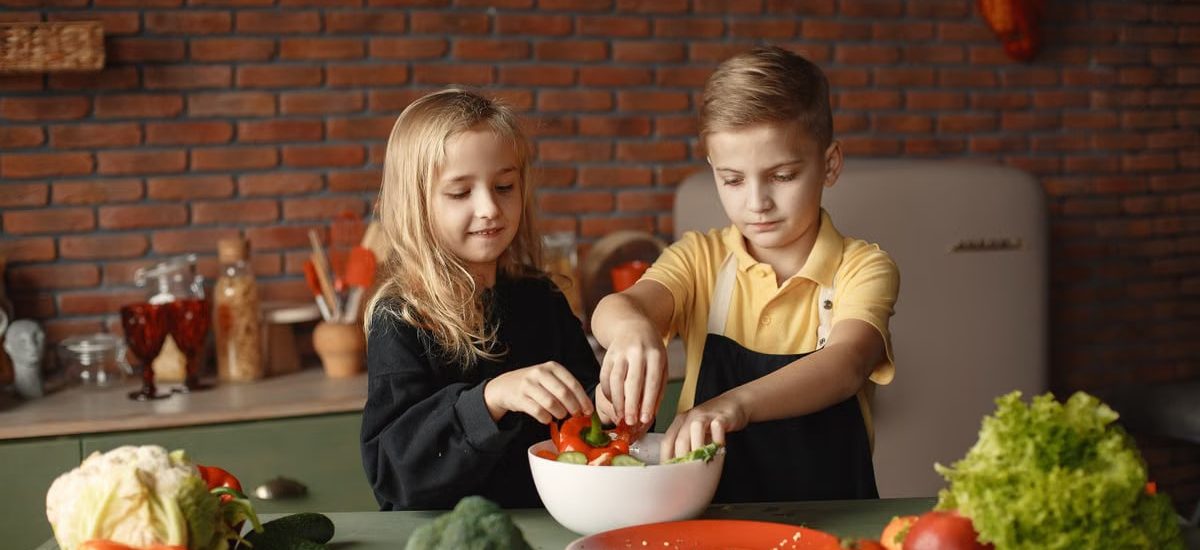Cheerful girl helping little boy in apron preparing salad while standing in kitchen near table with mix of fresh vegetables behind brick wall