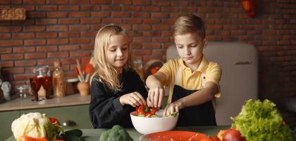 Cheerful girl helping little boy in apron preparing salad while standing in kitchen near table with mix of fresh vegetables behind brick wall