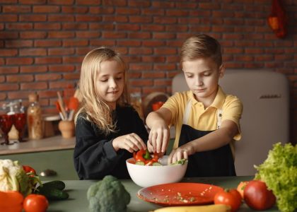 Cheerful girl helping little boy in apron preparing salad while standing in kitchen near table with mix of fresh vegetables behind brick wall